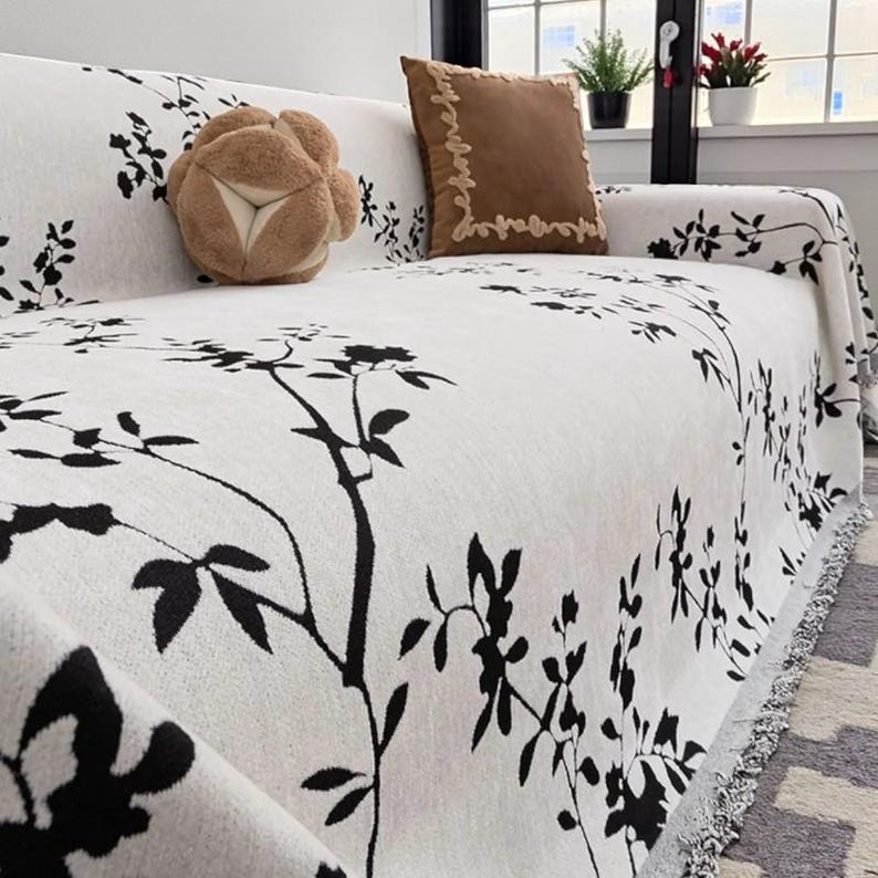 May include: A white sofa cover with a black floral pattern. The cover is draped over a sofa and features a fringe along the edge. Two decorative pillows in brown and tan hues are on the sofa.