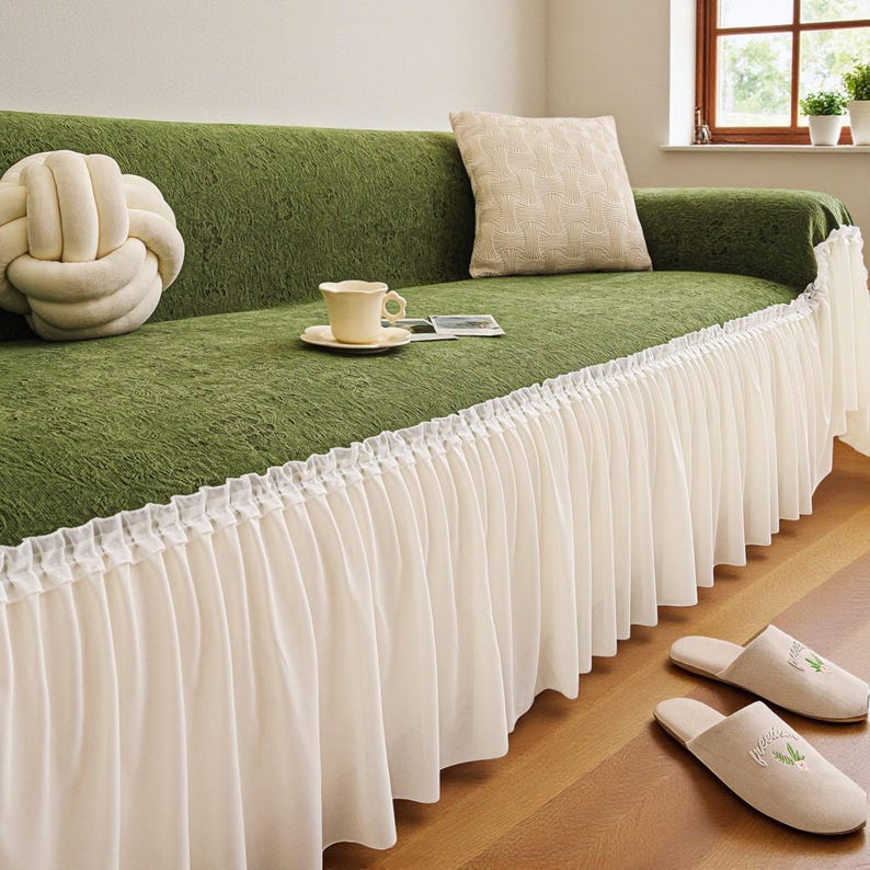 May include: A green sofa with a white skirt and matching throw pillows. A cream-colored cup and saucer sit on the sofa, along with a decorative knot pillow. Two pairs of beige slippers with green leaf embroidery are on the floor.