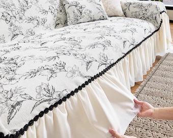May include: A white sofa cover with a black and gray floral pattern. The cover has a ruffled skirt with a black trim. Decorative pillows with floral patterns are on the sofa.