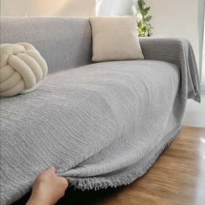 Puede incluir: Un sofá gris claro con una manta texturizada a juego y ribete de flecos. Dos cojines decorativos en crema y blanquecino están en el sofá. Una mano tira de la manta.