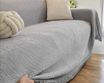 May include: A light gray sofa with a matching textured throw blanket and fringe trim. Two decorative pillows in cream and off-white are on the sofa. A hand is pulling up the blanket.