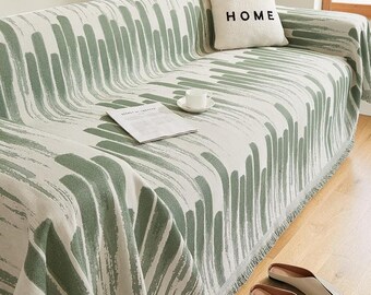 May include: A sofa covered in a cream-colored throw blanket featuring a sage green brushstroke pattern. A square throw pillow with the text LOVE HOME sits on the sofa, alongside a white ceramic cup on a saucer and a folded paper document.