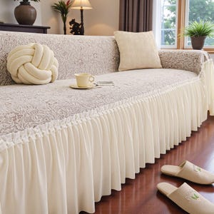 May include: A cream-colored sofa with a floral patterned cover and a pleated skirt. A decorative knot pillow and a square pillow rest on the sofa. A teacup and saucer sit on the sofa, and a pair of slippers are on the floor.