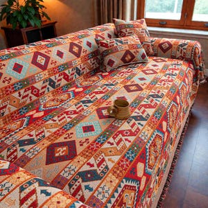 May include: A patterned sofa cover and cushions in shades of red, orange, blue, and cream. A brown mug is on the sofa. The cover has a fringed edge. The geometric design is eye-catching.