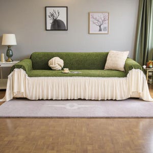 May include: A green sofa covered with a decorative slipcover. The slipcover has a ruffled cream-colored skirt and a matching pillow. A decorative knot pillow and a small teacup and saucer are on the sofa.