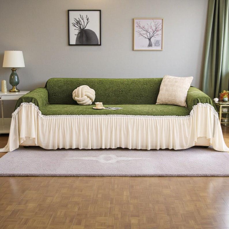 May include: A green sofa covered with a decorative slipcover. The slipcover has a ruffled cream-colored skirt and a matching pillow. A decorative knot pillow and a small teacup and saucer are on the sofa.