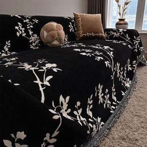 May include: Black sofa cover with a white floral pattern. The cover is draped over a sofa, with a fringed edge. Two throw pillows and a decorative ball are on the sofa.
