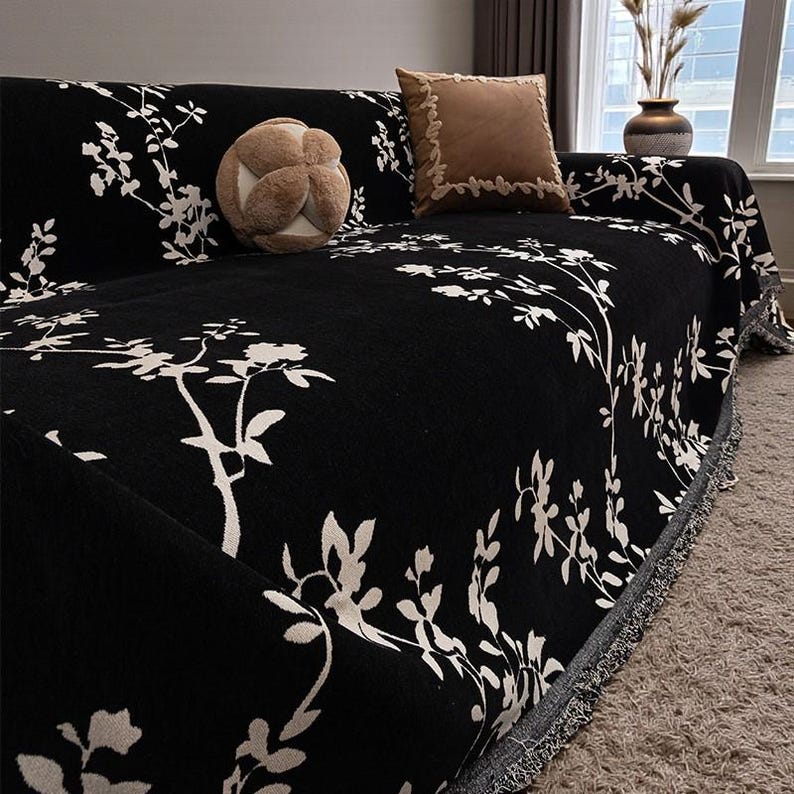 May include: Black sofa cover with a white floral pattern. The cover is draped over a sofa, with a fringed edge. Two throw pillows and a decorative ball are on the sofa.