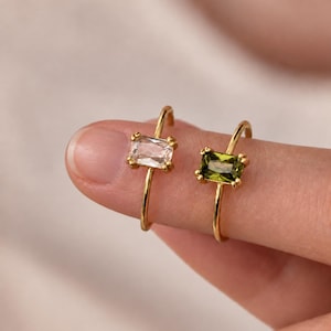 May include: Two gold rings with thin bands, each featuring a single rectangular baguette-cut gemstone. One ring has a clear, colorless stone, while the other ring features a deep olive green stone, both held in place by four small metal prongs.