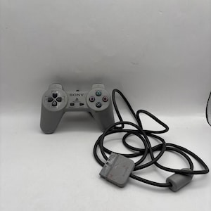 May include: A gray Sony PlayStation controller with a black cord. The controller has a directional pad, action buttons, and a PlayStation logo. The cord has a rectangular connector and a cylindrical end.
