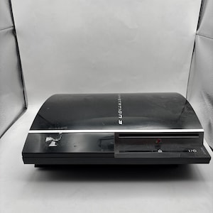 May include: A black Sony PlayStation 3 game console featuring a curved top surface with the text PlayStation 3 printed in white. The front panel includes a disc slot, a power button, and a small PlayStation logo emblem on the right side.