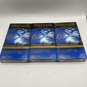 May include: Three Maxell VHS T-120 videocassette tapes. Each box is blue with the text "maxell" in gold, and "Premium Grade" in white. The boxes are designed for home theater applications and everyday recording.