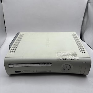 Könnte beinhalten: Eine weiße Xbox 360 Spielkonsole mit einem chromfarbenen Laufwerksschacht, einem Einschaltknopf und einem Lüftungsgitter auf der Oberseite. Ein rechteckiger Aufkleber mit dem Text ATTENTION ist vorne angebracht und beschreibt den Hardwarestatus.