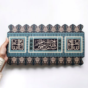 May include: A decorative wall hanging with a blue and beige patterned border and three rectangular panels with Arabic calligraphy in black and white. The panels are surrounded by a blue and white patterned border.