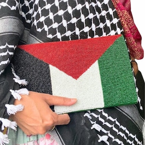 May include: A beaded clutch bag featuring the colours of the Palestinian flag: black, red, white and green. The bag is being held by a person wearing a black and white keffiyeh.