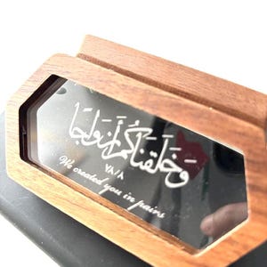 May include: A wooden box with a mirrored surface, inscribed with Arabic calligraphy and the English words "We created you in pairs." The box has a geometric design and a natural wood finish, with white text.