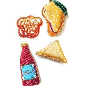 May include: Four embroidered felt food ornaments: a mango, a samosa, a bottle of red sauce with a blue label that says "Rooh Afza", and a swirl of orange and yellow.