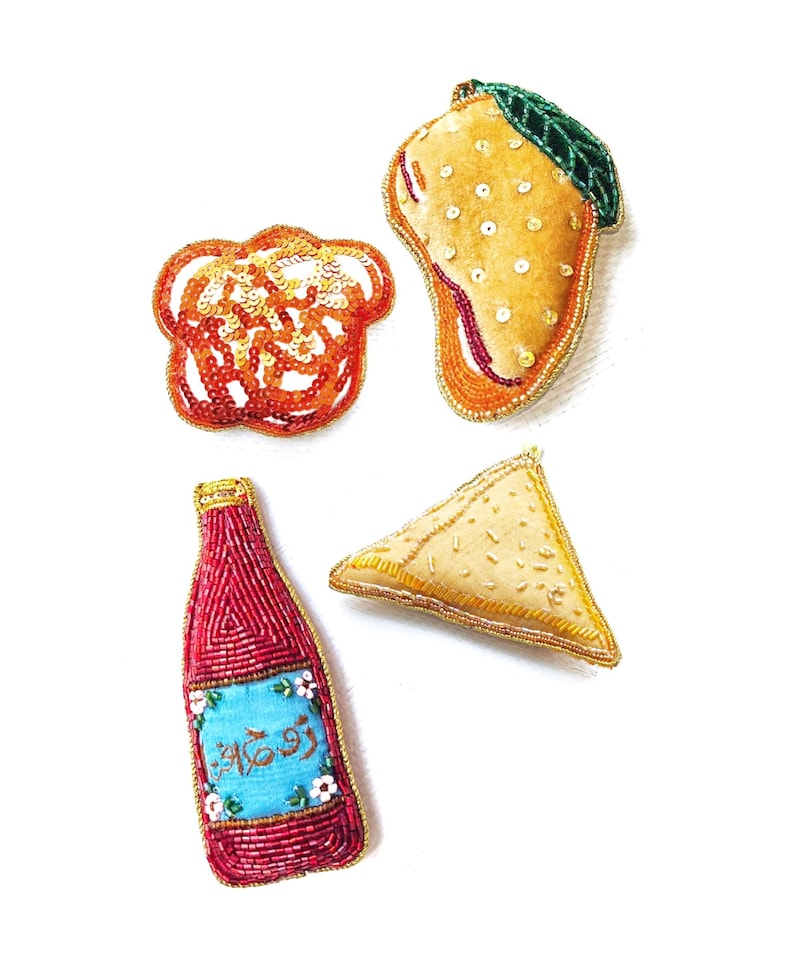 May include: Four embroidered felt food ornaments: a mango, a samosa, a bottle of red sauce with a blue label that says "Rooh Afza", and a swirl of orange and yellow.