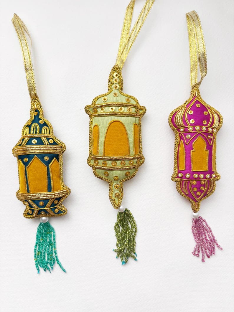 May include: Three decorative hanging lanterns with beaded tassels. The lanterns are made of fabric and embroidered with gold thread. The lanterns are in the colors of teal, green, and pink.