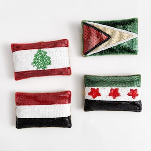 May include: Four beaded flag-themed rectangular patches. The flags represent Lebanon, Guyana, Yemen, and Syria. Each patch features a unique design with red, white, black, green, and gold beads. The patches are displayed on a white background.