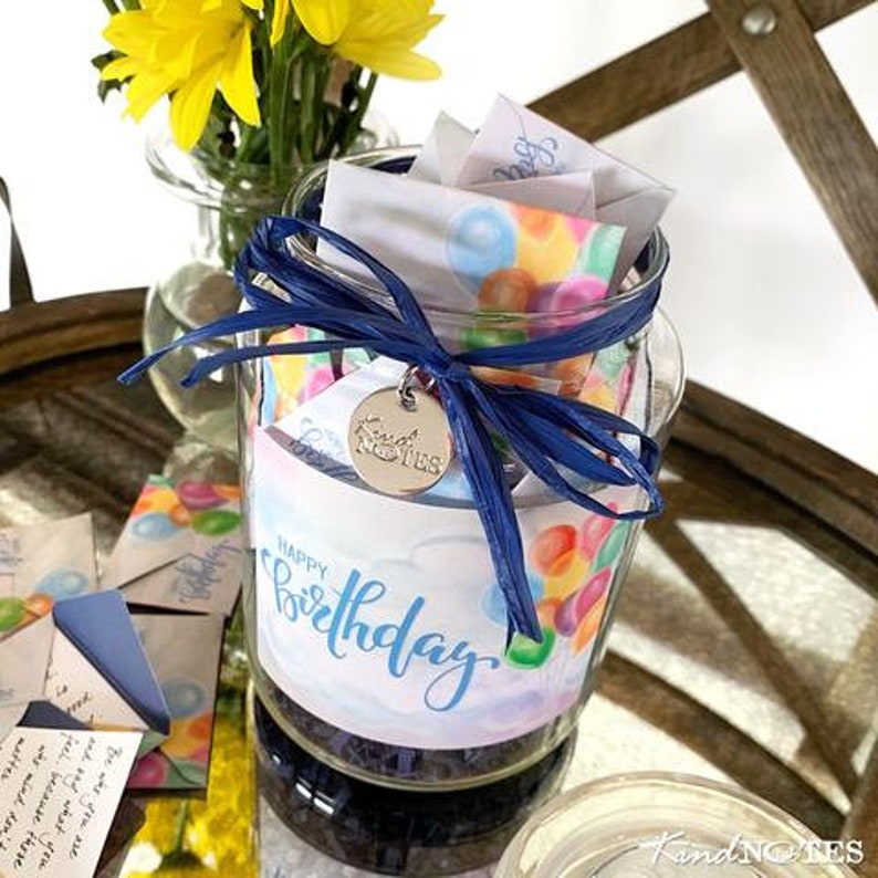 Op de afbeelding: Een glazen pot gevuld met kleurrijke papieren briefjes, vastgebonden met een blauw lint en een zilveren hanger met de tekst "Kind Notes". De pot heeft een label met de tekst "Happy Birthday".