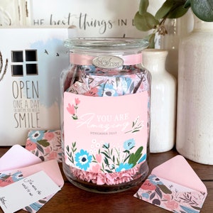 May include: A glass jar filled with pink and blue floral patterned paper notes. The jar has a pink ribbon and a silver label that says "Kind Notes". The jar has a pink label that says "You Are Amazing Remember That" with a floral design.