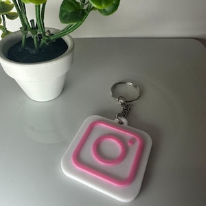 May include: White and pink Instagram logo keychain. The keychain features a white square with a pink Instagram logo in the center. The keychain is attached to a silver metal keyring.