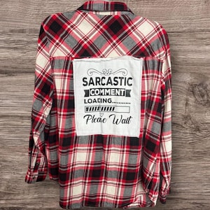 May include: Red, black, white, and gray plaid flannel shirt with a white patch on the back. The patch reads "Sarcastic Comment Loading... Please Wait" in black text. The shirt has long sleeves and a button-down front.