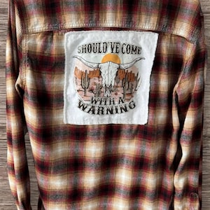 May include: A long-sleeved, plaid flannel shirt in shades of red, brown, and tan. The back features a white patch with a desert scene, a bull skull, and the text "SHOULD'VE COME WITH A WARNING".
