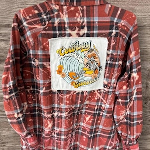 May include: Red and black plaid button-up shirt with a white patch on the back. The patch features the words "Cowboy Summer" with a skeleton riding a surfboard. The shirt has a distressed, bleached design.