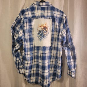 May include: Blue and white plaid flannel shirt with a skull and sunflower graphic on the back. The graphic is on a white patch and features a skull with sunflowers and other floral elements in shades of orange, yellow, and blue.