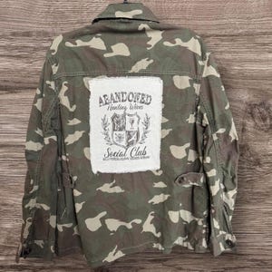 May include: A camouflage print utility jacket featuring a large, rectangular white fabric patch sewn onto the back. The patch displays the text "ABANDONED Hunting Wives Social Club" and "HELLO HUNTING SEASON, GOODBYE HUSBAND" with a crest graphic.