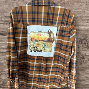 May include: A long-sleeve button-down shirt in a brown, navy blue, and white plaid pattern. The back features a large, rectangular white patch with a desert landscape graphic, a lasso, and the text "KEEP IT WILD" printed in dark brown letters.