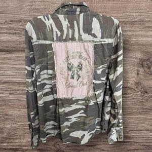 May include: A long-sleeve button-down shirt featuring a camouflage pattern in shades of green, brown, and gray. A rectangular light pink patch is sewn onto the back, displaying a silver bow graphic and the text Deserted Hunters' Wives Social Club.