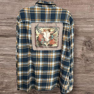 May include: A long-sleeved, button-down flannel shirt in a navy blue, yellow, and white plaid pattern. The back features a rectangular patch with a bull skull design, accented with floral elements. The shirt is a casual, Western-inspired garment.