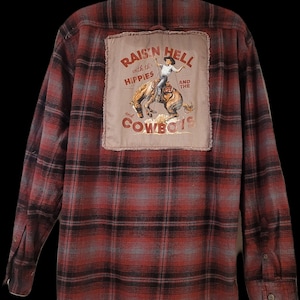 May include: A long-sleeve button-down shirt in a red, black, and gray plaid pattern. The back features a large, rectangular, frayed-edge tan patch with a graphic of a cowboy on a bucking horse and the text RAIS'N HELL with the HIPPIES AND THE COWBOYS.