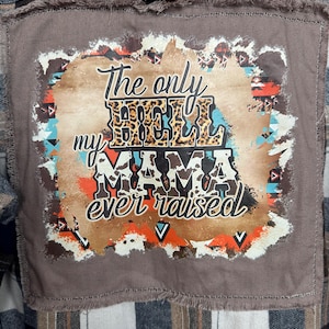 May include: A brown and black plaid flannel shirt with a distressed patch. The patch features the text "The only hood my mama ever raised" in a mix of fonts and leopard print.