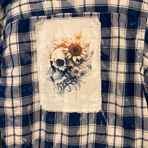 May include: A blue and white plaid button-down shirt featuring a rectangular white fabric patch sewn onto the back. The patch displays a detailed illustration of a human skull surrounded by sunflowers with blue and orange watercolor accents.