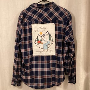 May include: A long-sleeve button-down shirt in a navy blue, white, and red plaid pattern. The back features a rectangular white fabric patch with a graphic of mountains, a tent, a campfire, and a river, with the text Making Memories one campsite at a time.