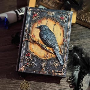 Puede incluir: Un diario decorativo en relieve con un cuervo posado en una rama. La portada presenta un diseño detallado de cuervo tridimensional con bayas rojas y detalles dorados. El diario tiene una estética oscura y gótica.