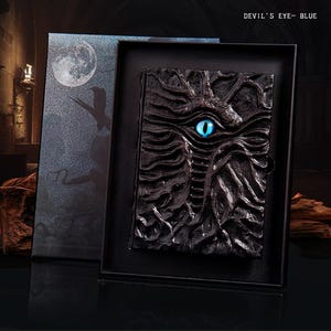 May include: A black, textured journal with a blue eye design, presented in a black box. The journal cover features a detailed, carved appearance. The box has a moon and silhouette design. Text on the box reads "DEVIL'S EYE - BLUE".