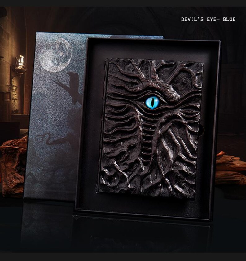 May include: A black, textured journal with a blue eye design, presented in a black box. The journal cover features a detailed, carved appearance. The box has a moon and silhouette design. Text on the box reads "DEVIL'S EYE - BLUE".