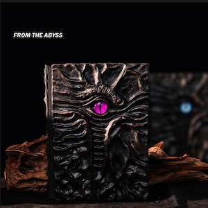 May include: A dark, textured book cover with a prominent, glowing purple eye design. The cover is intricately carved with a detailed, organic pattern. The text "FROM THE ABYSS" is displayed at the top. The book's spine is visible, and a second book is blurred in the background.