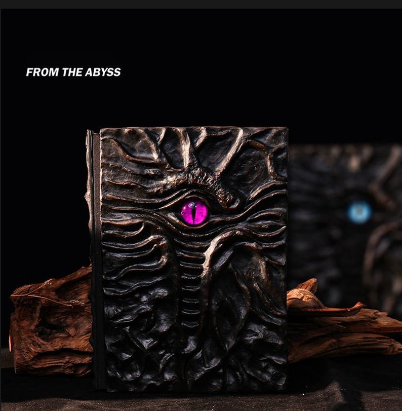 May include: A dark, textured book cover with a prominent, glowing purple eye design. The cover is intricately carved with a detailed, organic pattern. The text "FROM THE ABYSS" is displayed at the top. The book's spine is visible, and a second book is blurred in the background.