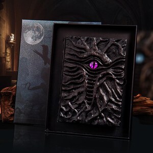 May include: A black, intricately designed journal with a dragon motif, featuring a prominent purple eye. The journal is presented in a matching black box with a moon and silhouette design. The cover has a textured, carved appearance.