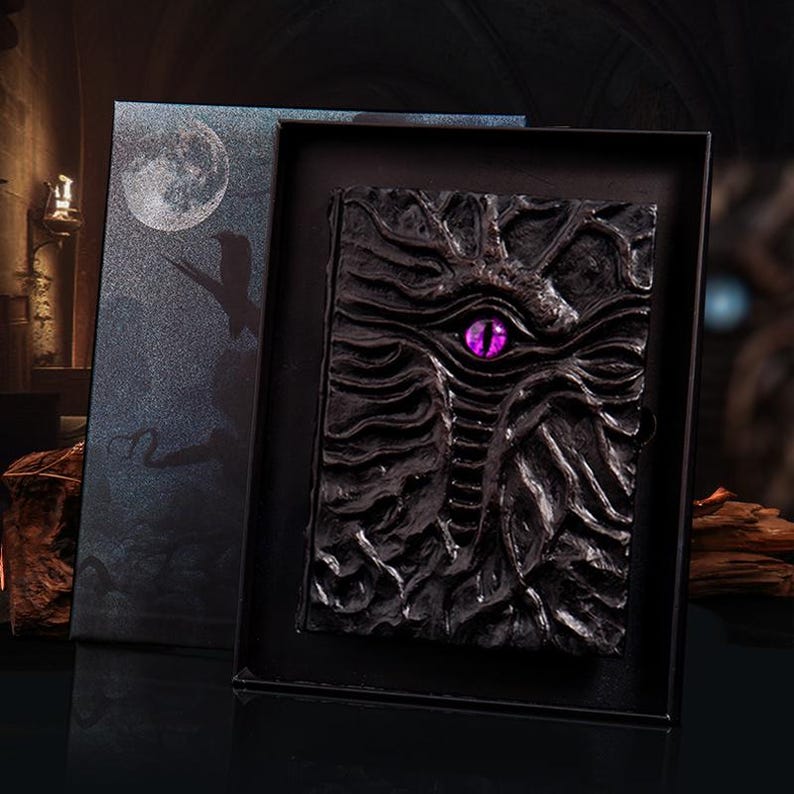 May include: A black, intricately designed journal with a dragon motif, featuring a prominent purple eye. The journal is presented in a matching black box with a moon and silhouette design. The cover has a textured, carved appearance.