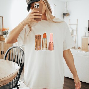 May include: A cream-coloured t-shirt with a watercolour illustration of three beauty products: a lipstick, a nail polish, and a lipstick. The lipstick is orange, the nail polish is pink, and the lipstick is gold. The person is wearing a black baseball cap.