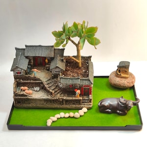 May include: A miniature Asian-style village planter with a green succulent plant. The planter features a detailed model of a traditional village with miniature figures, pigs, and a water buffalo figurine on a green base.