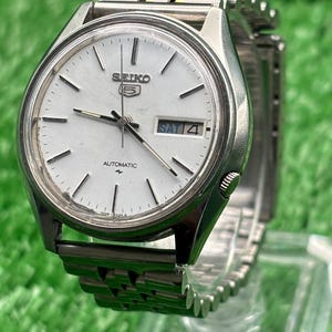 Op de afbeelding: Een zilveren Seiko 5 automatische polshorloge met een witte wijzerplaat. Het horloge heeft een dag- en datumweergave, zilveren wijzers en een zilveren band. Het horloge wordt getoond op een transparante standaard tegen een groene achtergrond.