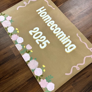 May include: A brown paper sign with the words "Homecoming 2025" in white and yellow lettering. The sign is decorated with pink flowers and green leaves along the bottom left side and pink ribbon accents.